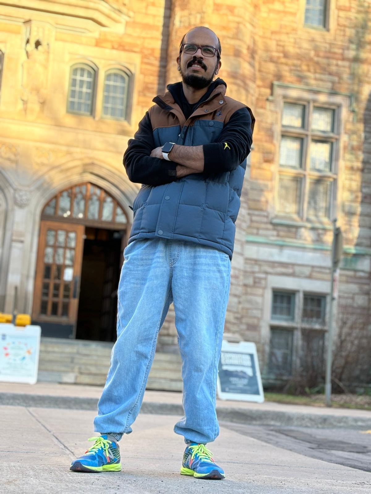 Suleman is standing confidently with his arms crossed in front of a historic building. He is wearing a blue and brown puffer vest over a black hoodie, light blue jeans, and colorful sneakers with bright green laces. The building behind him has a stone facade with arched windows and a wooden door. He has a beard and glasses, and he is wearing a smartwatch on his left wrist. The photo is taken from a low angle, giving him a towering presence.