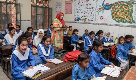 The image shows a classroom in Pakistan with young girls in blue school uniforms sitting at wooden desks, engaged in their studies. A female teacher, wearing a traditional Pakistani dress with a headscarf, is standing while holding a book. The classroom walls are decorated with colorful educational charts and Urdu calligraphy, enhancing the learning environment. The students appear attentive, with some reading aloud and others following along in their textbooks.