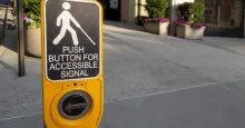 A yellow accessible pedestrian crossing button on a sidewalk, featuring a sign with a person using a cane and the text 'Push button for accessible signal.' In the background, there are potted plants with green shrubs and pink flowers near a building entrance.