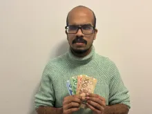 Suleman is standing against a plain off-white wall. He is wearing a light green knitted turtleneck sweater and holding several Canadian banknotes in his hands, including $100, $50, $20, and $5 bills. His expression is serious, and he is looking directly at the camera.