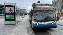 A Société de transport de Montréal (STM) bus, number 28-049, is parked at a bus stop in Montreal. The bus stop has a shelter with an advertisement for Fizz, a telecommunications company. A metro sign in the background suggests proximity to a metro station. The surrounding environment includes urban buildings, a restaurant, and pedestrians walking on the sidewalk under an overcast sky.