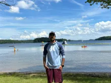 Suleman wearing sunglasses, a cap, and a blue Hurley hoodie stands on the grassy shore of a lake with a calm expression. Behind him, people are enjoying the water, some wading near the shore while others kayak or use floating platforms. A blue and yellow inflatable structure is visible in the distance, along with lush green trees surrounding the lake under a bright blue sky with scattered white clouds.