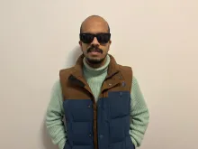 Suleman is wearing black sunglasses, a light green knitted turtleneck sweater, and a blue and brown puffer vest. He stands against a plain off-white wall with a relaxed posture, hands in his pockets, and a slight smile on his face.