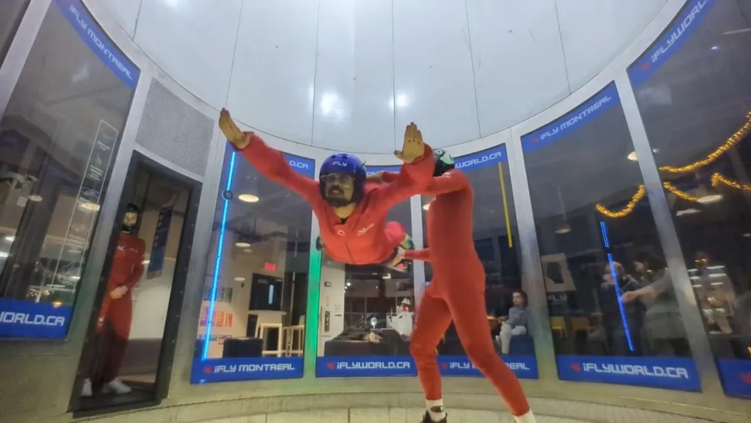 This image Suleman experiencing indoor skydiving at iFLY Montreal. He is wearing a red flight suit and a blue helmet while floating in a vertical wind tunnel. An instructor in a similar red suit is assisting him. The background features the transparent walls of the wind tunnel, with onlookers watching from outside. The iFLY Montreal branding is visible on the glass panels.