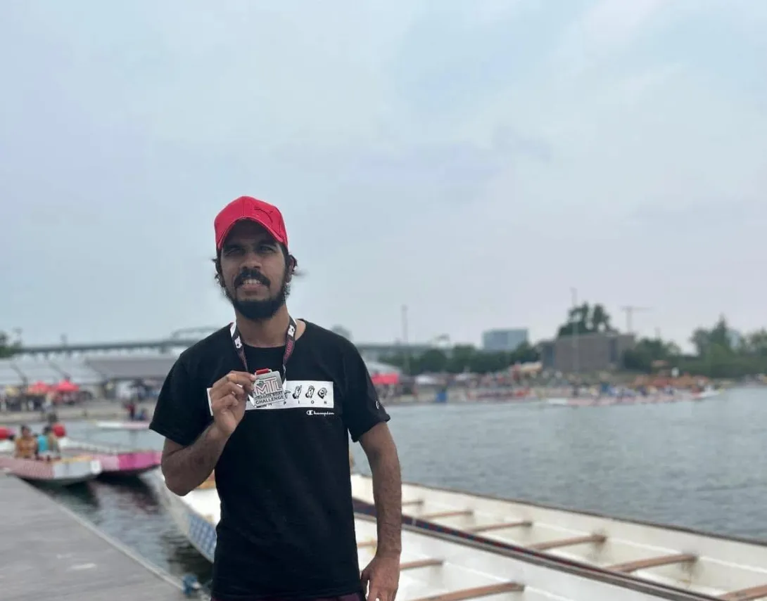 Suleman is standing near a waterfront, at a dragon boat event. He is wearing a black t-shirt and a red cap while holding a medal in his hand. In the background, there are boats, water, and a crowd of people who seem to be engaged in a competitive activity.