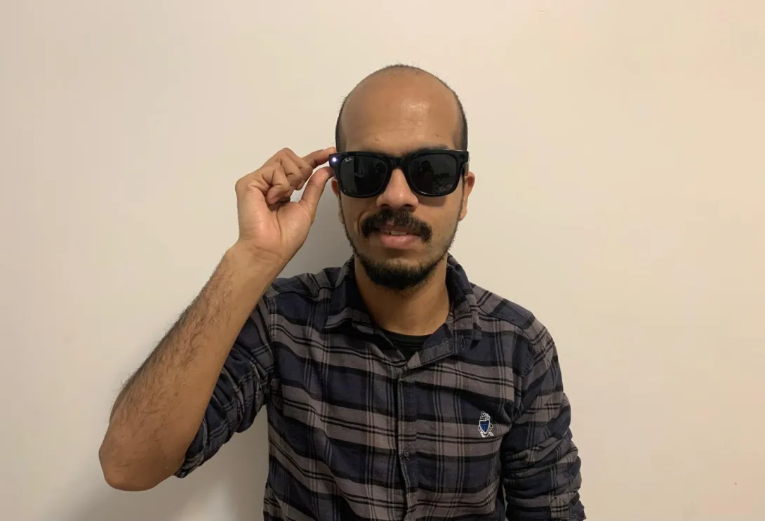 Suleman, wearing a black and grey plaid shirt and Ray-Ban Meta smart glasses, stands against a plain white wall. He has a beard and mustache and is smiling slightly. He is adjusting the glasses with his right hand while a small LED light is visible on the frame.