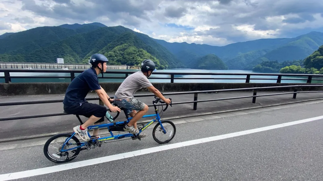 The image shows two people riding a tandem bicycle along a scenic road near a body of water. They are wearing helmets and cycling on a paved road with a railing separating them from the lake or river. The background features lush green mountains under a partly cloudy sky, creating a peaceful and adventurous atmosphere.