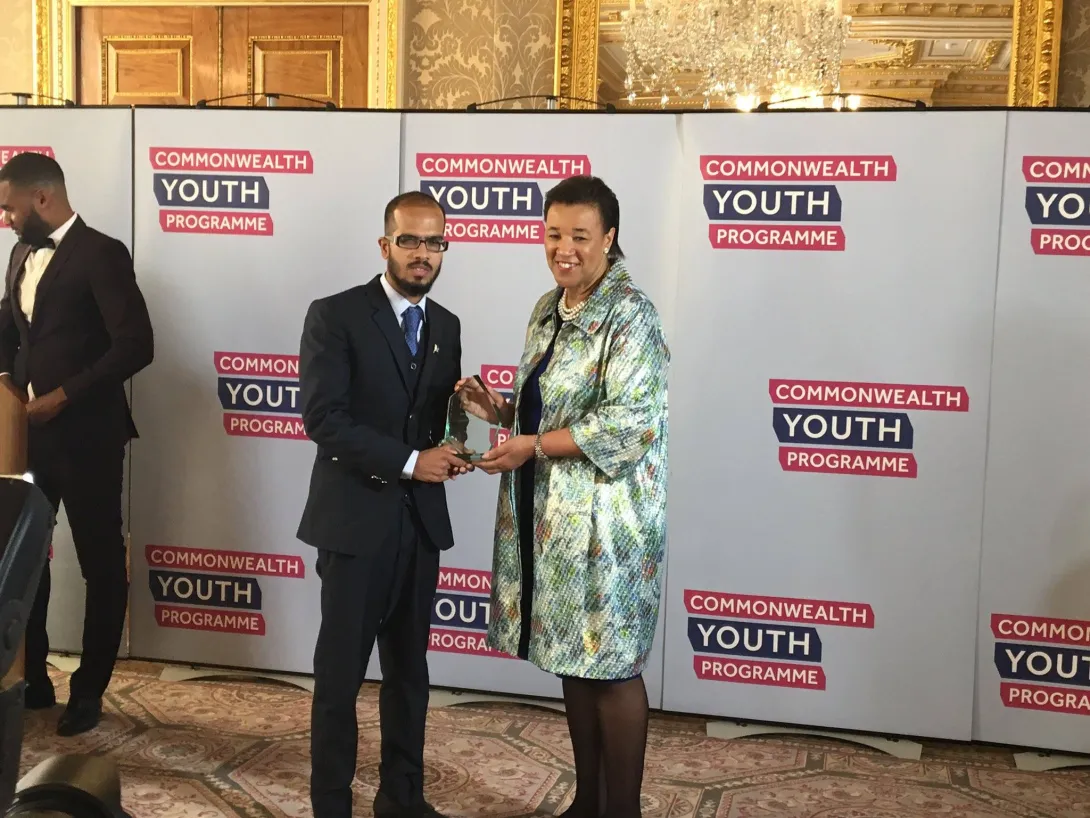 The image captures Suleman Arshad from Pakistan receiving an award from Commonwealth Secretary-General Patricia Scotland at a Commonwealth Youth Programme event.