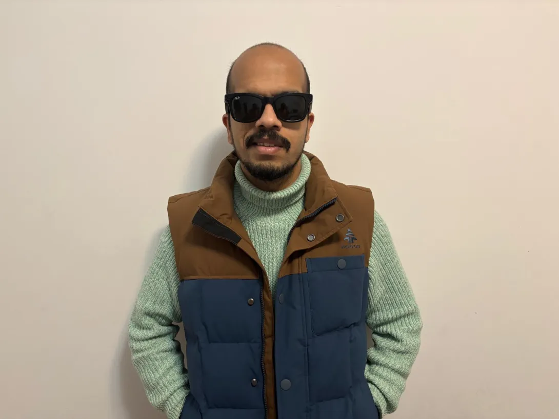 Suleman is wearing black sunglasses, a light green knitted turtleneck sweater, and a blue and brown puffer vest. He stands against a plain off-white wall with a relaxed posture, hands in his pockets, and a slight smile on his face.